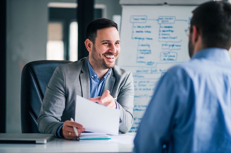 how-to-hire-employees-1 A male hiring manager who understands how to hire employees meets with a male candidate.