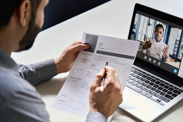 A bearded male hiring manager interviews a Black male candidate during a virtual meeting, while reviewing his CV