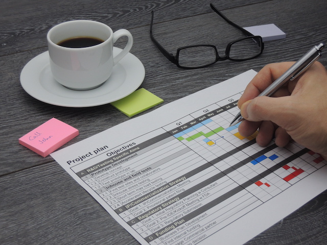 Someone writes on a project plan printout on a desk near a cup of coffee and pair of glasses