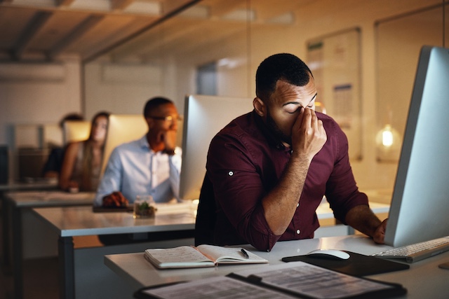 A stressed and tired male employee shuts his eyes at his desk late at night