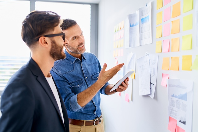 A manager helps his male employee with a project plan posted on a wall
