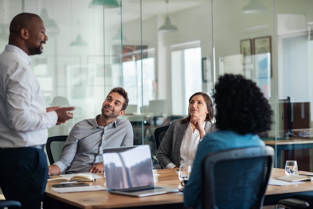 A Black male executive stands before his team, leading a meeting