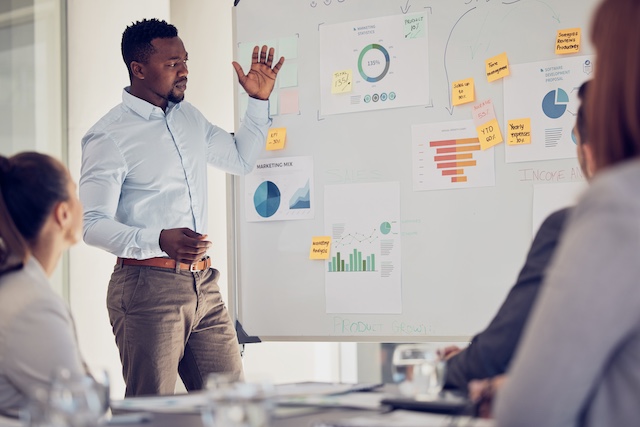 A Black male leader presents a plan on a whiteboard to his team
