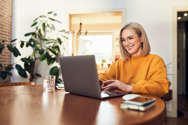 Happy woman with glasses uses laptop at dining table for her outplacement program, demonstrating why companies offer outplacement